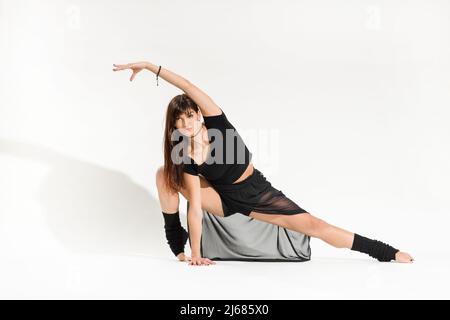 Full body of young female dancer in black outfit performing front stretch with flexed instep and raised arm on white background Stock Photo