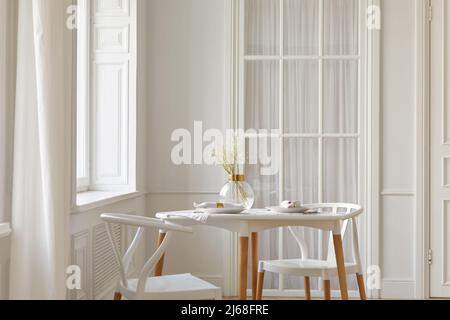 Glass vase with dried flowers and dishware placed on table near chairs and window in white spacious room at home Stock Photo