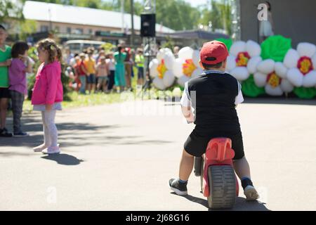 Children on a walk in the park. A boy on a red toy motorcycle in black sneakers, shorts, vest, white T-shirt, red cap. Behind the girl in pink and whi Stock Photo