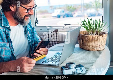 Digital nomad and remote worker freedom lifestyle concept. Adult man buying on line using laptop and mobile phone connection inside a camper van in tr Stock Photo