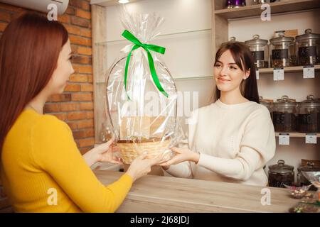 seller sells gift set, festive packaging. woman buys basket with gift in tea shop. Private medium business, individual entrepreneur. smiling, happy, satisfied Stock Photo