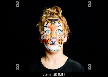 Portrait of a boy with strange and scary gesture, made up with tiger face, isolated on black background. Stock Photo