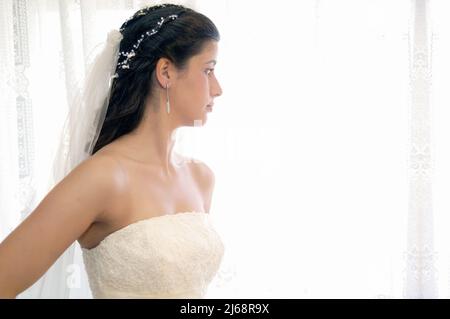 Pretty girl dressed in the wedding dress standing by a window with white curtains looking outside pensively on her wedding day. High key image. Stock Photo