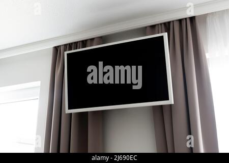 Empty TV screen in a hotel hanging on the wall. Bedroom television as design mockup template in a motel. Curtains and windows next to the display. Stock Photo