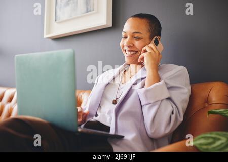 Portrait of multiracial LGBTQ mid adult woman using smartphone and laptop on couch and concentrating Stock Photo