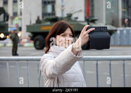 Happy girl taking selfie pictures on smartphone on background of armored vehicles of russian military forces during a rehearsal of Victory Day parade Stock Photo