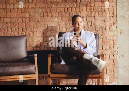 Portrait of confident multiracial LGBTQ mid adult woman sitting on retro chair and using smartphone in front of exposed brick wall Stock Photo