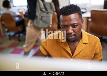 A Black man concentrates on his desktop computer while colleagues walk past Stock Photo