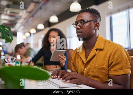 A man sits at his desk in an open plan office typing and looking at his screen Stock Photo