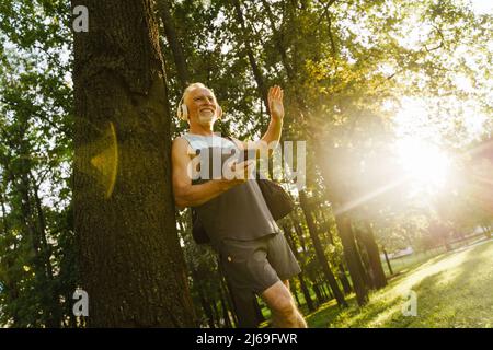 Grey senior man smiling and waving hand while using cellphone in park Stock Photo