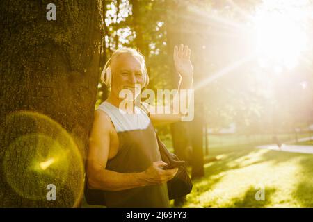 Grey senior man smiling and waving hand while using cellphone in park Stock Photo