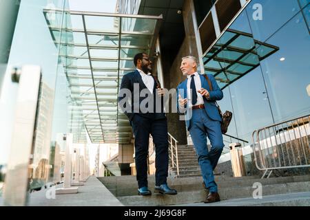 Multiracial men dressed in suits talking and gesturing while going down stairs outdoors Stock Photo