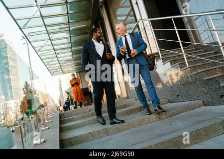 Multiracial men dressed in suits talking and gesturing while going down stairs outdoors Stock Photo