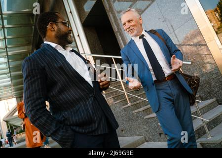 Multiracial men dressed in suits talking and gesturing while going down stairs outdoors Stock Photo