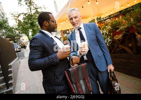 Multiracial two men wearing suits talking while drinking coffee outdoors Stock Photo