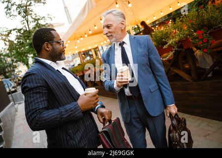Multiracial two men wearing suits talking while drinking coffee outdoors Stock Photo
