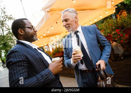 Multiracial two men wearing suits talking while drinking coffee outdoors Stock Photo