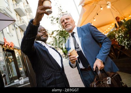 Multiracial two men wearing suits talking while drinking coffee outdoors Stock Photo