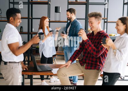 Front view of five cheerful young business multiracial startup colleagues having fun enjoying communication during coffee break in co-working space. Stock Photo