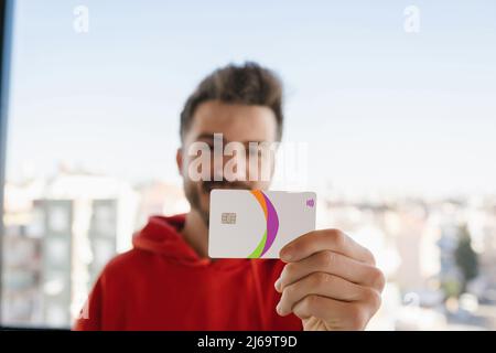 Happy man wearing red hoodie holding credit card with a happy face standing and smiling with a confident smile. Selective focus on credit card or debi Stock Photo