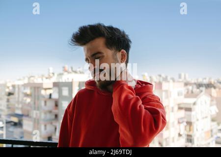 Feeling exhausted. Unhappy man wearing red hoodie suffering from neck pain at balcony. Frustrated young handsome man looking exhausted while leaning m Stock Photo