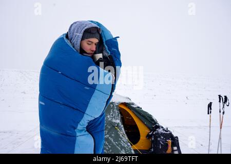 A guy in a sleeping bag is standing on a snow near the tent and drinking coffee Stock Photo