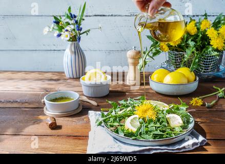 Dandelion salad with olive oil, lemon juice and spices on brown wooden table. Human hand pouring olive oil to dish. Stock Photo