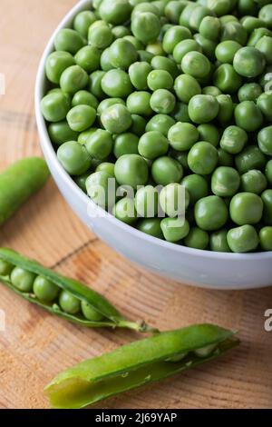 Green peas in white bowl with fresh pods on the wooden background. Stock Photo