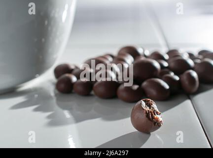 chocolate covered raisins, hazelnuts, coffee beans, almonds pouring out of a white bowl on a white table lit by the sun lighting through the window Stock Photo