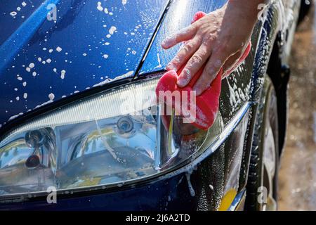 Man is washing car outside. Hand cleaning car in garage in front of house. Male hand holding pink sponge with foam. Stock Photo