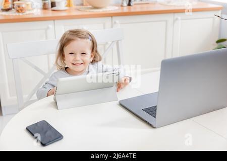 Happy little girl smiling and looking to the camera while watching cartoons. Pretty kid sitting at home while quarantine. The concept of learning at Stock Photo