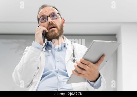 Male physician with stethoscope sitting at desk, talking on mobile phone and looking at digital tablet, discussing a patient diagnostic. High quality photography Stock Photo