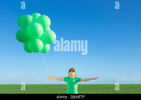 Happy child playing with bright multicolor balloons outdoor. Kid having fun in green spring field against blue sky background. Healthy and active life Stock Photo