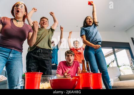 friends jumping for joy watching e-sports on TV after their team's victory. group of young watching soccer on tv. Stock Photo