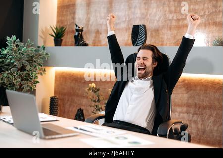 Success, joy, victory concept. Caucasian happy businessman in a formal stylish suit, sit in his modern office, rejoicing at a great deal, financial profit, successful project, gesturing with his hands Stock Photo