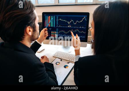 Successful caucasian colleagues, cryptocurrency traders, man and woman, sit at a table in a modern office, looking at computer screen, work with stock charts and documents, analyze the growth dynamics Stock Photo