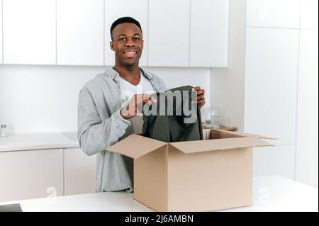 Happy cheerful african american guy unpacks his long awaited package, examines the clothes that he bought online, stands in kitchen, satisfied with the purchase. Online shopping, delivery concept Stock Photo