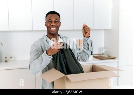 Smiling attractive black man unpacked his parcel, happy about getting a long expected order. Handsome guy shopping in internet stores, buying new clothes online, online shopping, delivery concept Stock Photo