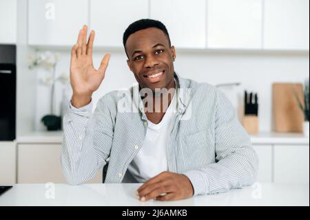 Online video chat. Photo of a positive african american man, sits at home, looks at web camera talks through video call, male freelancer or mentor communicates to client remotely, greeting, smiling Stock Photo