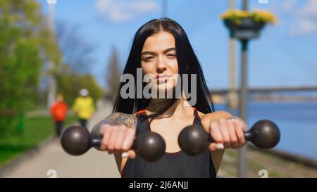 Serious brunette woman holds two old dumbbells during sunny day and looks at you Stock Photo