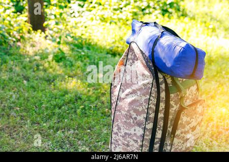 Defocus military backpack and blue tent or sleeping bag. Army bag on green grass background near tree. Military camouflage army rucksack. Tourist summ Stock Photo