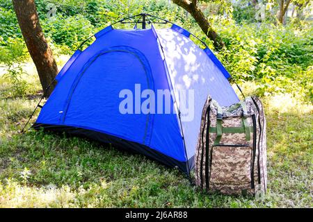 Defocus blue open tourist tent standing on green nature background. Army backpack. Tourism concept. Summer vacation in forest, camping. Lifestyle. Out Stock Photo