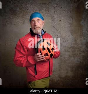 portrait of smiling senior male cyclist in bright red biking jacket and skull cap holding a helmet against a grunge concrete wall Stock Photo