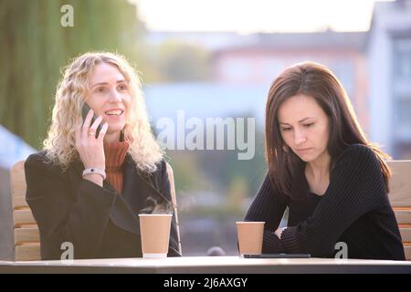 Unhappy young woman waiting angrily while her friend is talking happily on sellphone with someone else and ignoring her. Friendship problems concept. Stock Photo