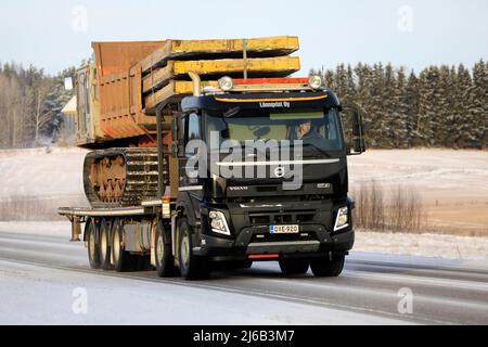 Black Volvo FMX truck Lonnqvist Oy hauls Hitachi CG 110 tracked dumper on flatbed trailer on highway 52 in winter. Salo, Finland. December 28, 2021. Stock Photo