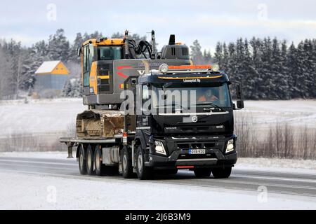 Black Volvo FMX truck Lonnqvist Oy hauls Volvo excavator on flatbed trailer along highway 52 on a day of winter. Salo, Finland. December 27, 2021. Stock Photo