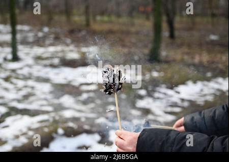 An adult woman's hand holds a burning paper flower in front of a winter park. The black charred paper is smoking. Stock Photo