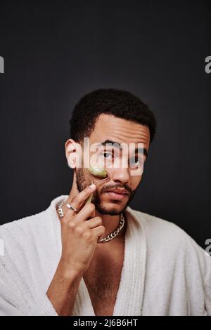 Portrait of young handsome man in bathrobe making facial massage with mesoroller against the black background Stock Photo