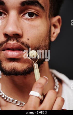 Close-up of handsome mixed race man massaging face with stone jade roller and looking at camera Stock Photo
