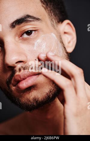 Close-up of bearded young man applying face cream on his face and looking at camera Stock Photo